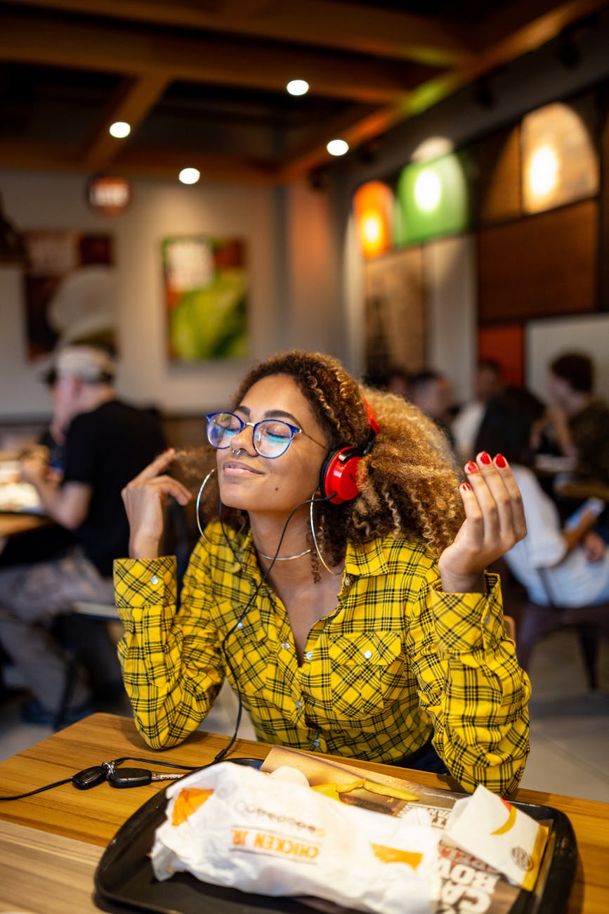 Image of a Black woman with curly hair wearing a yellow plaid shirt, listening to music on headphones while sitting in a restaurant.