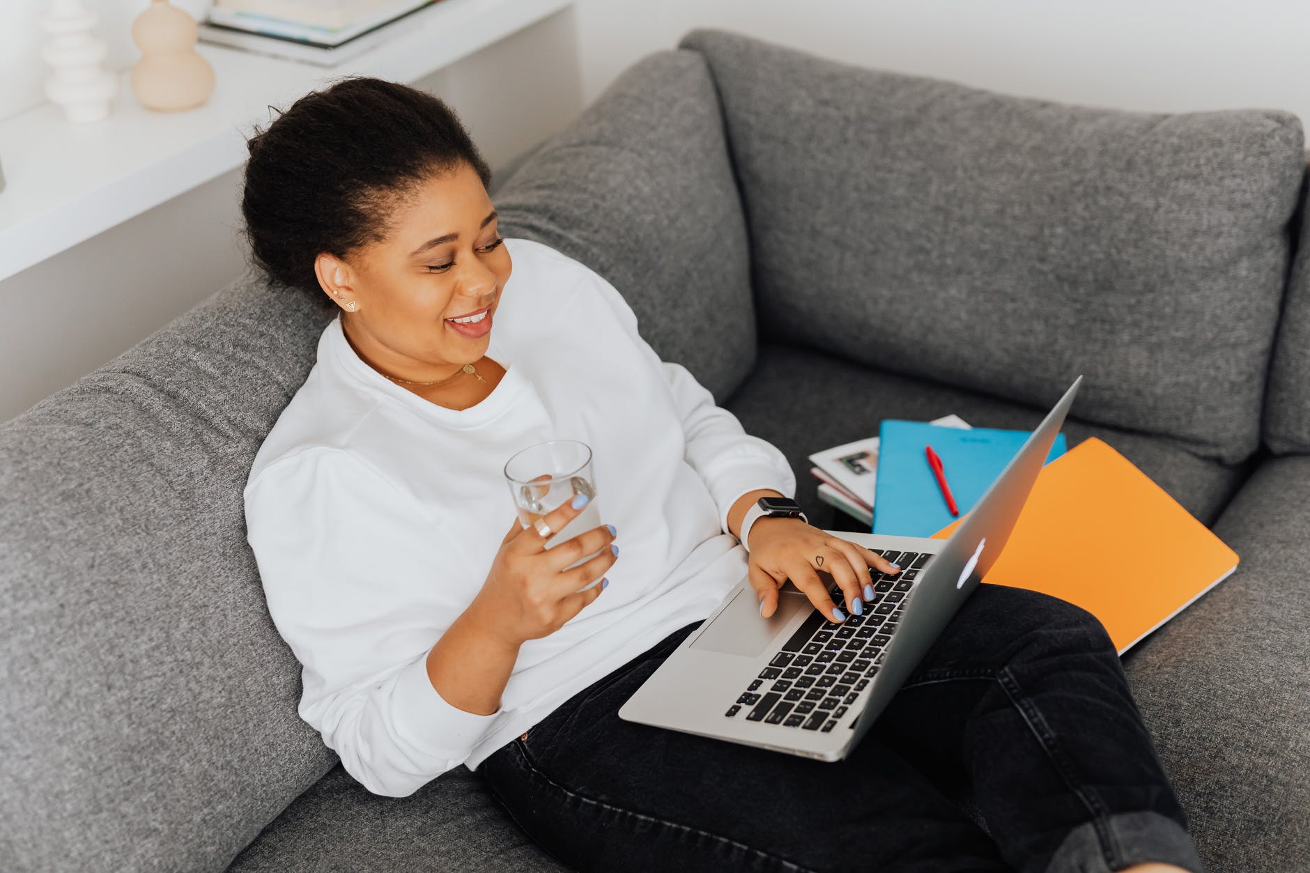 Image of a Black woman with short hair dressed in a white sweatshirt and black pants. She has a glass of water in hand, several books to the side of her and a laptop on her lap as she sits on a gray couch.