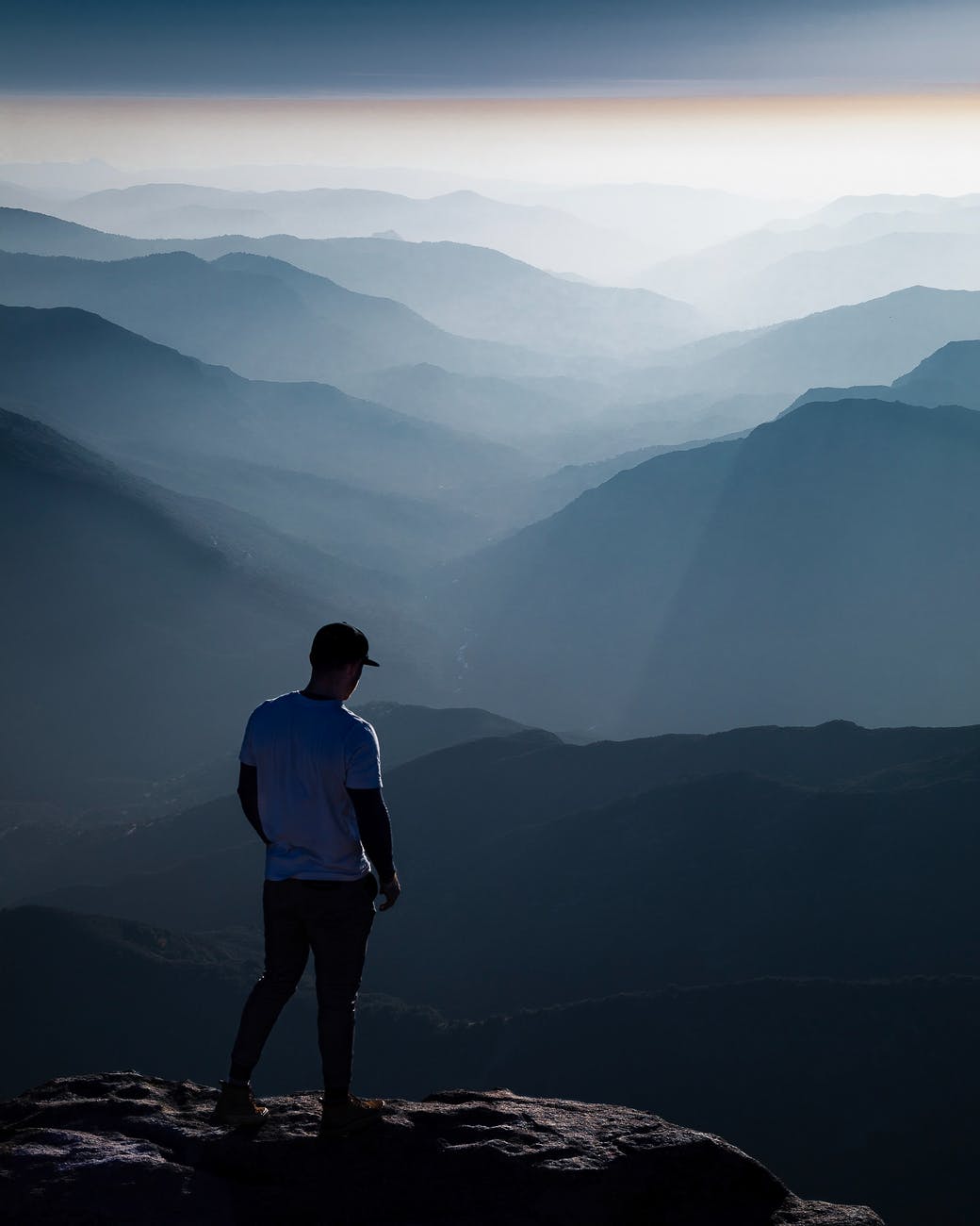Image of a person on top of a mountain looking out into the distance with many mountain tops ahead of them. They are being kissed by the sunlight.